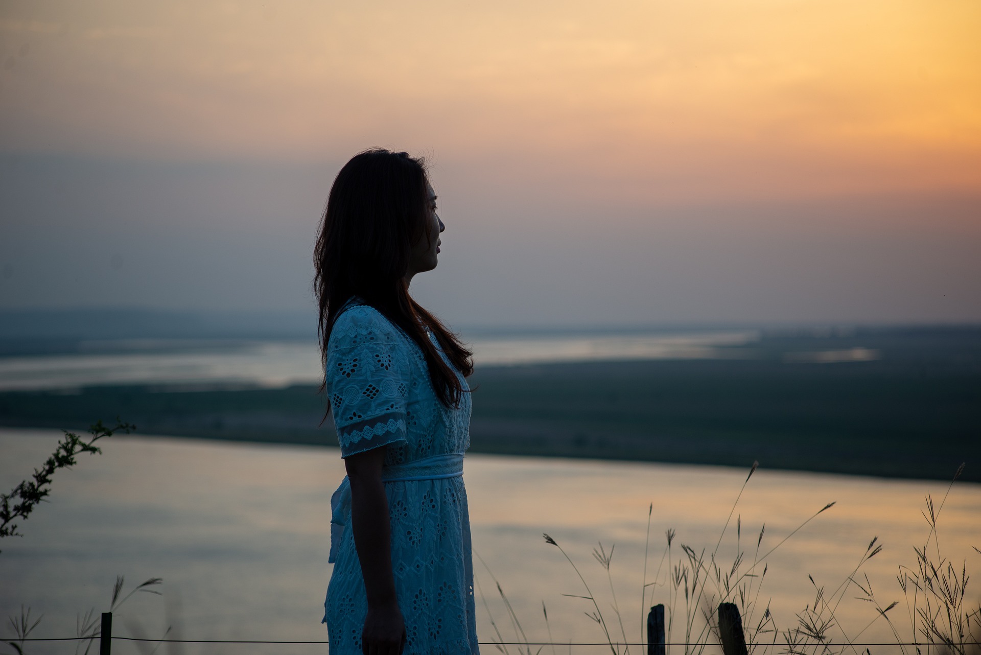 Woman standing in profile looking into the distance over water and land at sunset, representing reflection and tranquility