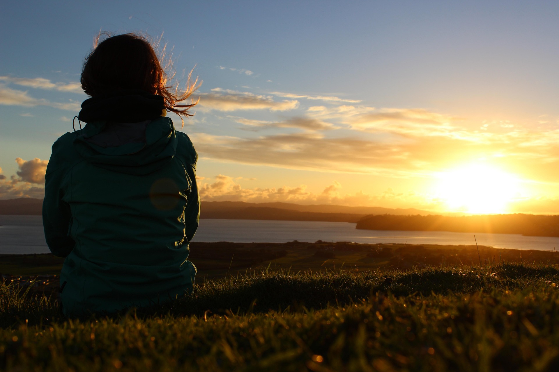 Woman sitting on grass facing the sea at sunset, reflecting in a calm and peaceful moment