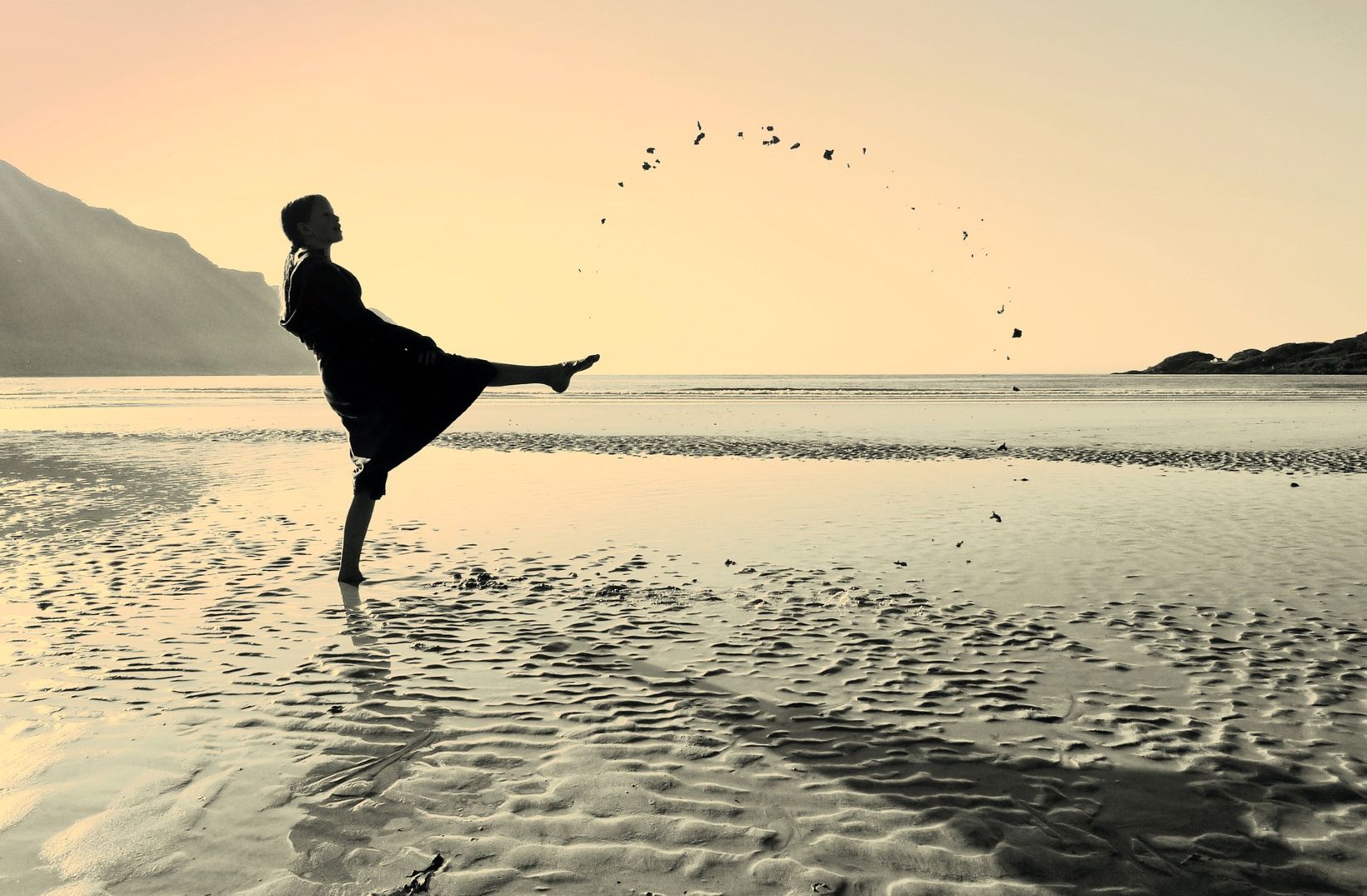 Woman standing in shallow water kicking water, with soft pink sky in black and white image, representing calm and inner peace