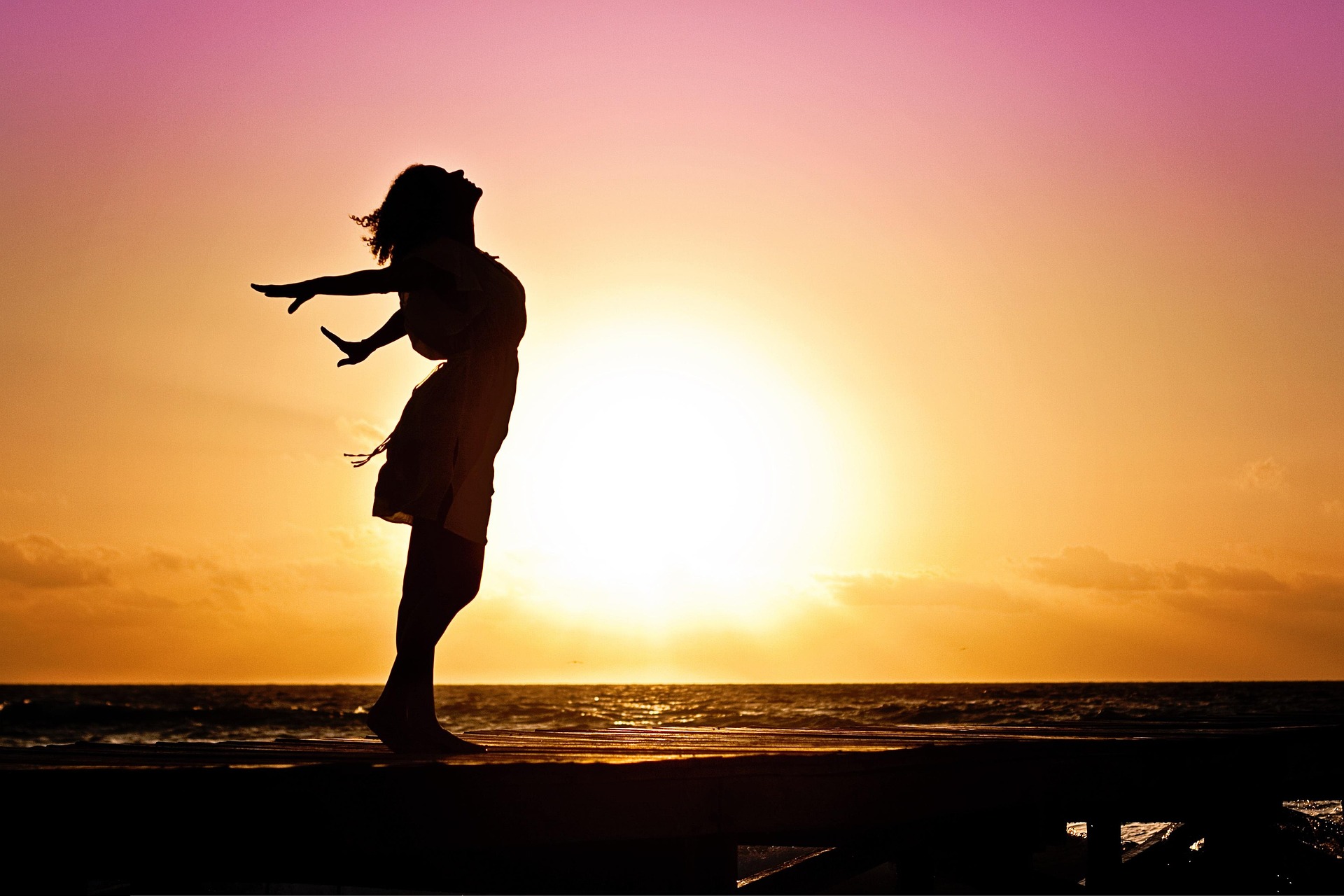 Silhouetted woman with arms open by the ocean at sunset, representing freedom and emotional openness